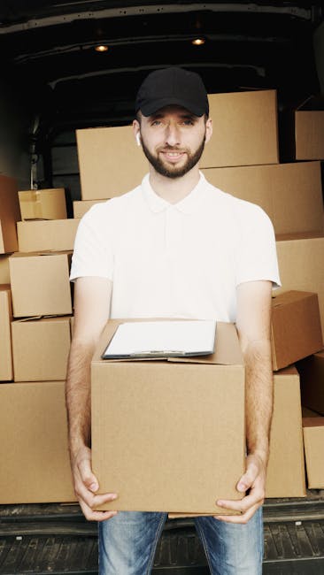 A man seated on a wooden chair outside a white van parked on a city street during a home relocation or moving process, with the van's rear doors open revealing packed cardboard boxes, wooden furniture
