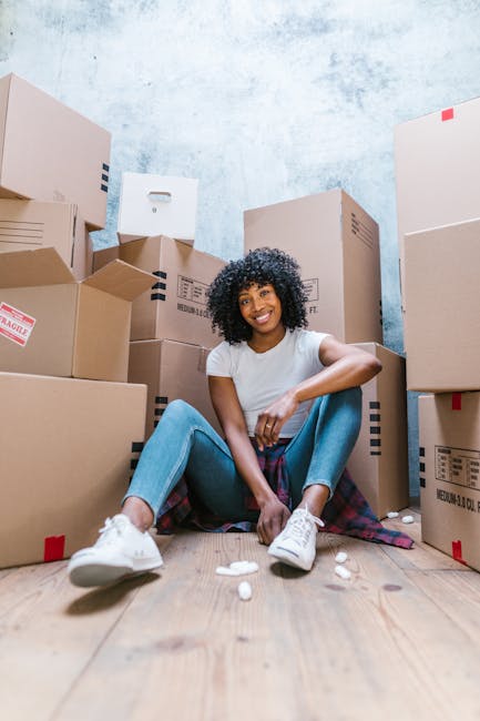 A young woman with curly dark hair and a bright smile is sitting on a wooden floor surrounded by numerous cardboard boxes of various sizes, some with labels and tape, in a room with a textured light blue wall. She is wearing a white t-shirt, light blue jeans, and white sneakers, and has a plaid shirt tied around her waist. In front of her are some packing peanuts, and she appears relaxed during a home relocation or packing process. The scene suggests an active moving or furniture transport operation, with the boxes likely prepared for a house move, reflecting components of professional removals by Removal Van Bow near Bow Road Station E3.