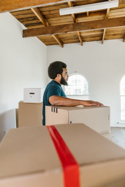 A man with dark curly hair and a beard, wearing a blue shirt, is seen inside a spacious room with white walls and large arched windows that let in natural light. He is standing behind several large cardboard boxes stacked on top of each other, with some boxes partially covered with red packing tape and plastic wrap, indicating they are packed for moving. The room has an open ceiling with exposed wooden beams and a wooden panel ceiling, suggesting it may be a converted loft or industrial-style property. The man appears to be in the process of preparing or organizing furniture and packing materials for a house removal. The scene is part of a home relocation, with the focus on furniture transport and packing process facilitated by [COMPANY_NAME] as part of their removals service. In the background, there are additional boxes and possibly small furniture items, all arranged on the floor close to the windows, which provide bright, diffused daylight that highlights the scene.