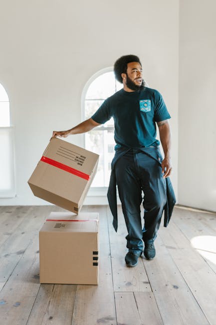 A young woman with curly dark hair and a bright smile is sitting on a wooden floor surrounded by numerous cardboard boxes of various sizes, some with labels and tape, in a room with a textured light blue wall. She is wearing a white t-shirt, light blue jeans, and white sneakers, and has a plaid shirt tied around her waist. In front of her are some packing peanuts, and she appears relaxed during a home relocation or packing process. The scene suggests an active moving or furniture transport operation, with the boxes likely prepared for a house move, reflecting components of professional removals by Removal Van Bow near Bow Road Station E3.