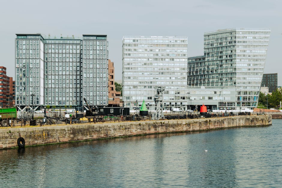 A view of a canal lined with modern, multi-storey residential buildings constructed with beige brick and dark paneling, featuring balconies and large windows. A metal railing runs along the water's ed
